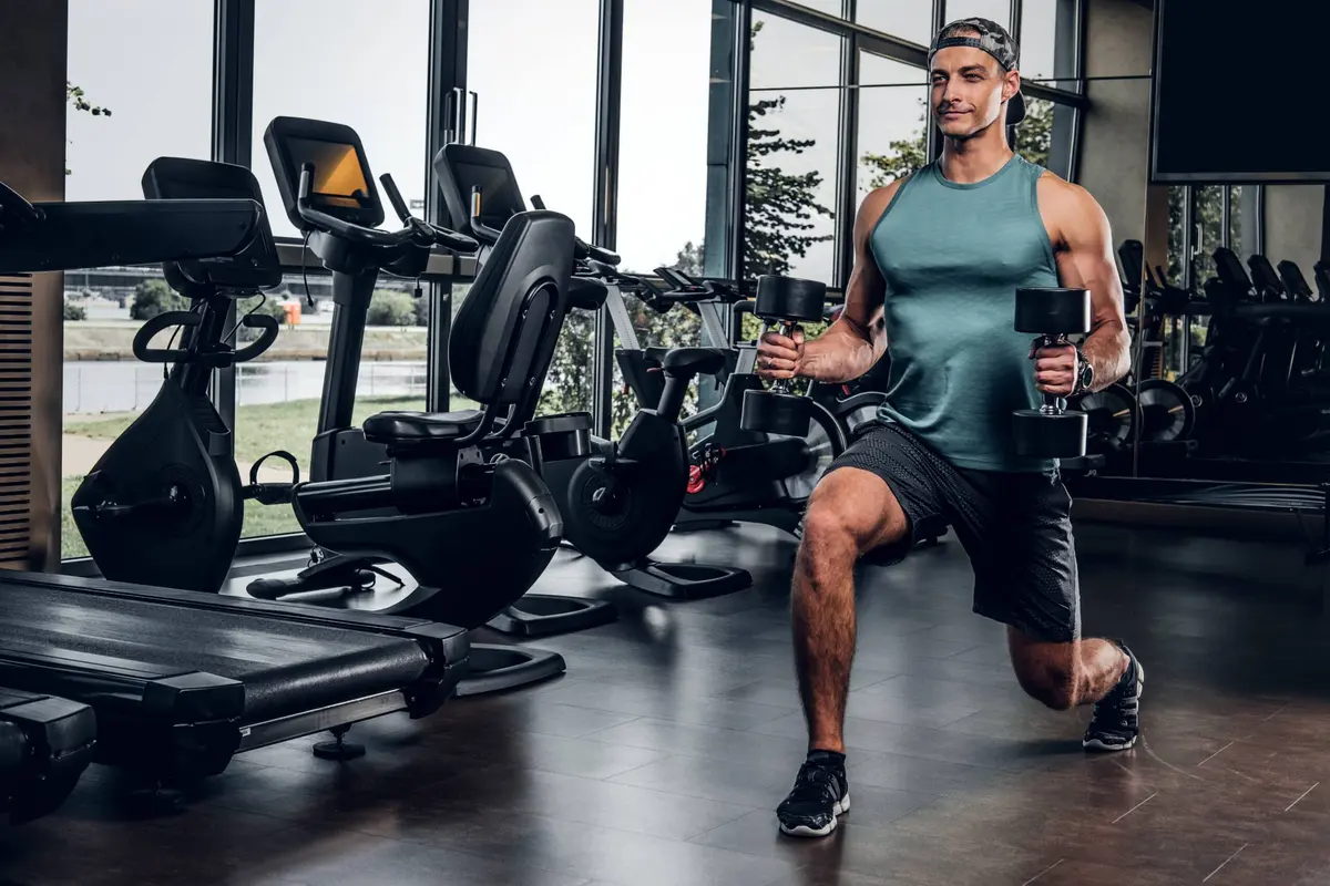 homem fazendo afundo com halteres na academia