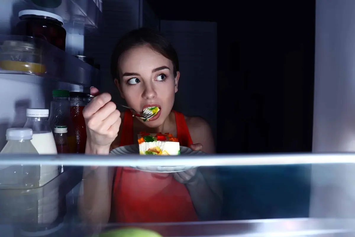 mulher comendo na frente da geladeira à noite