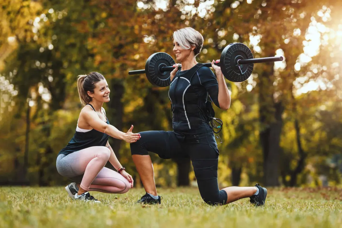personal trainer acompanhando mulher no afundo com barra ao ar livre