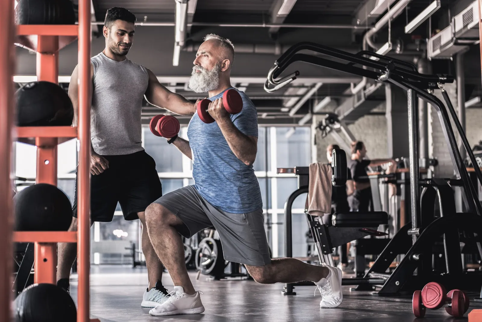 Personal trainer acompanhando um aluno idoso no exercício de avanço com halteres