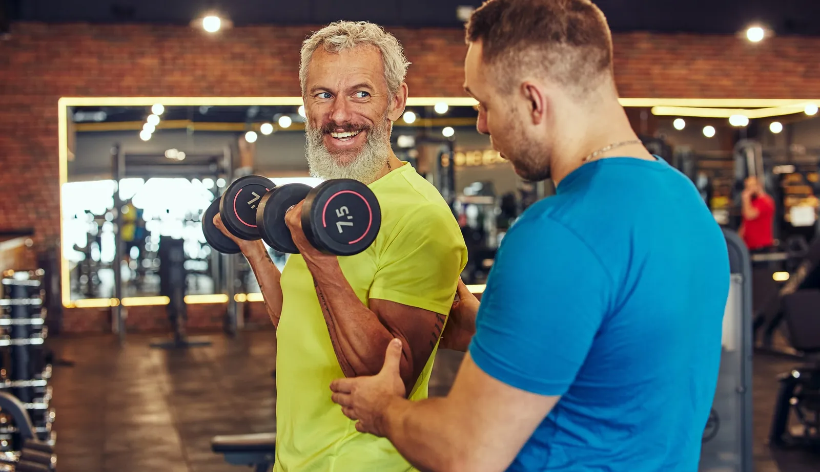 Personal trainer acompanhando um aluno idoso sorridente no exercício com halteres