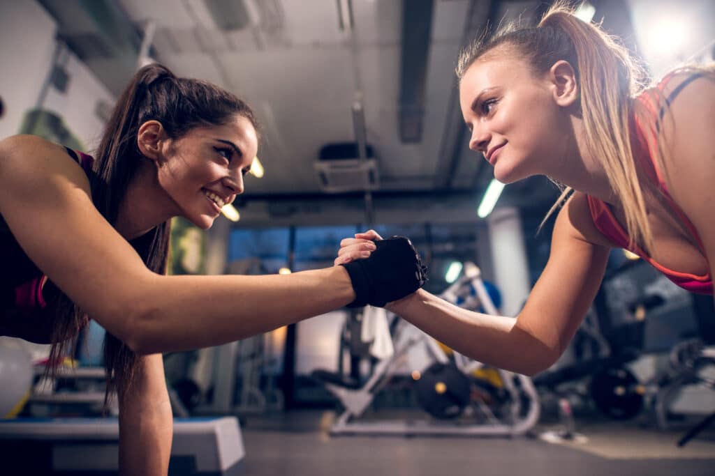 Two young motivated smiling attractive focused sporty active girls while doing push ups and holding hands together in the modern gym. amigas no treino amizade amigos