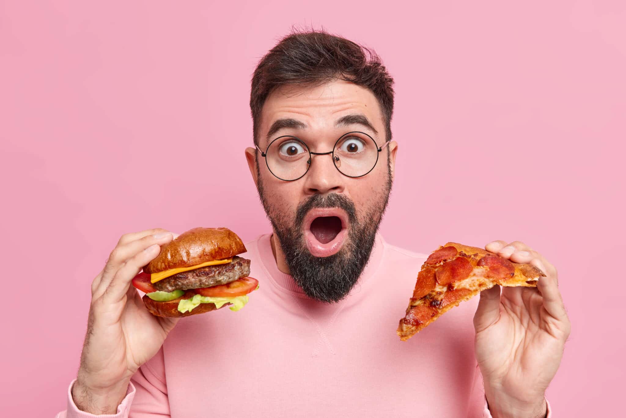 substituir fast food por alimentos saudáveis receitas saudáveis Surprised bearded man holds piece of pizza and hamburger eats junk food shocked to consume much calories wears round spectacles casual jumper isolated over pink background. Cheat meal concept